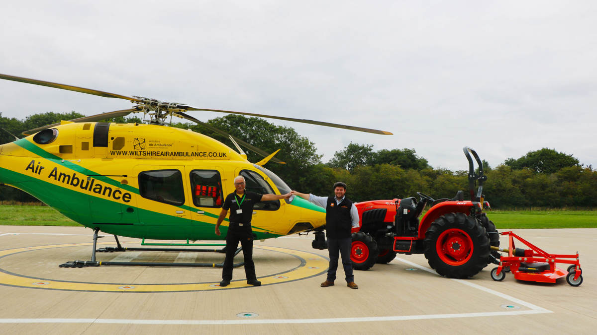 An orange Kubota tractor and trailer parked on the helipad next to the helicopter. There is a staff member from WAA and Howard & Sons handing over the keys.