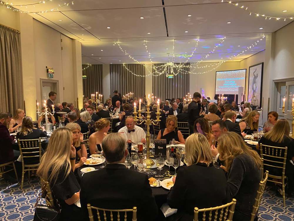 Black and gold themed charity ball. Groups of people wearing smart clothing, sitting on gold chairs around round tables