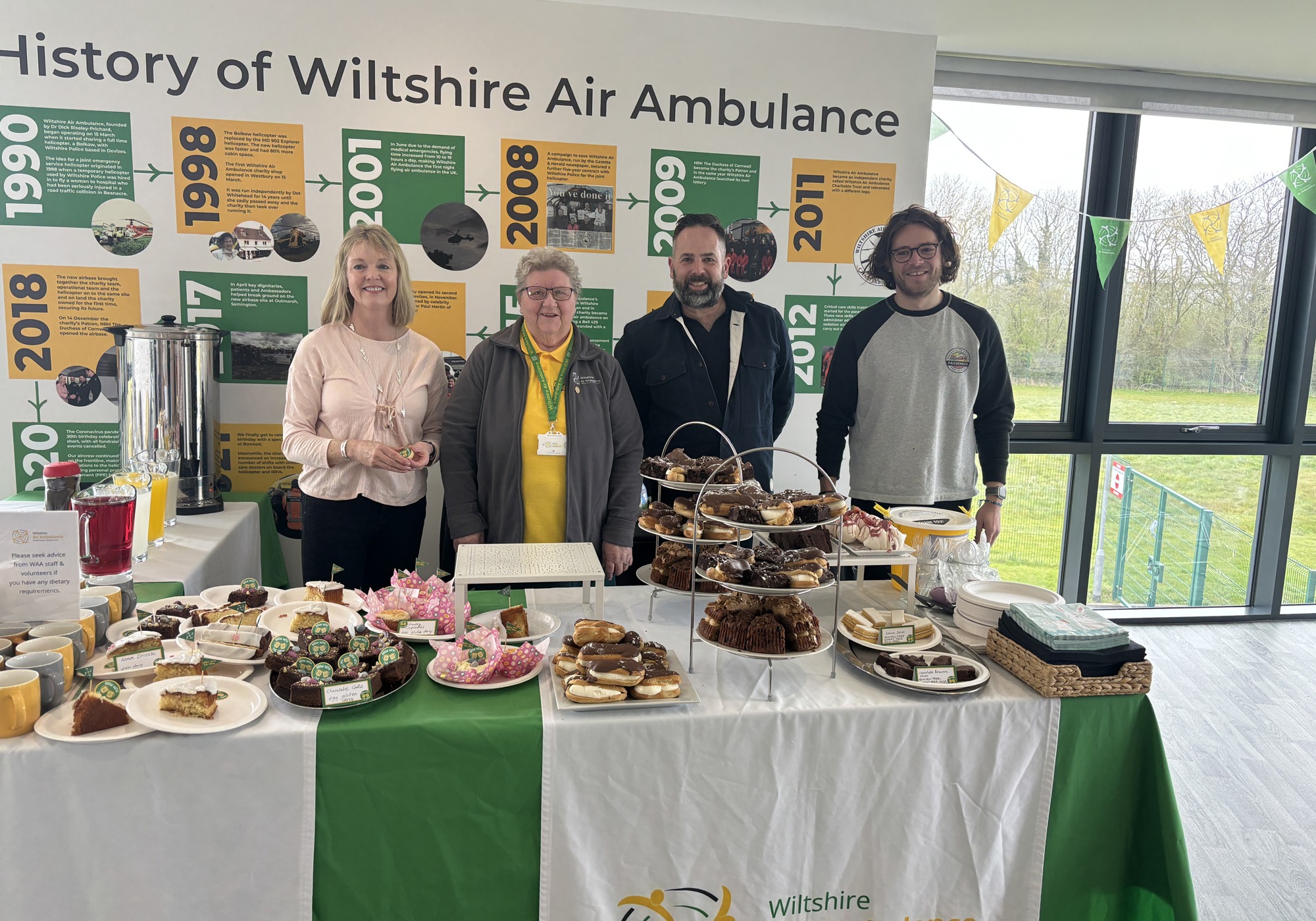 A table full of cakes, with four adults standing behind it, smiling at the camera A table full of cakes, with four adults standing behind it, smiling at the camera