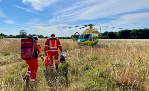 A photo of two paramedics, carrying kit bags, walking through long grass to the helicopter