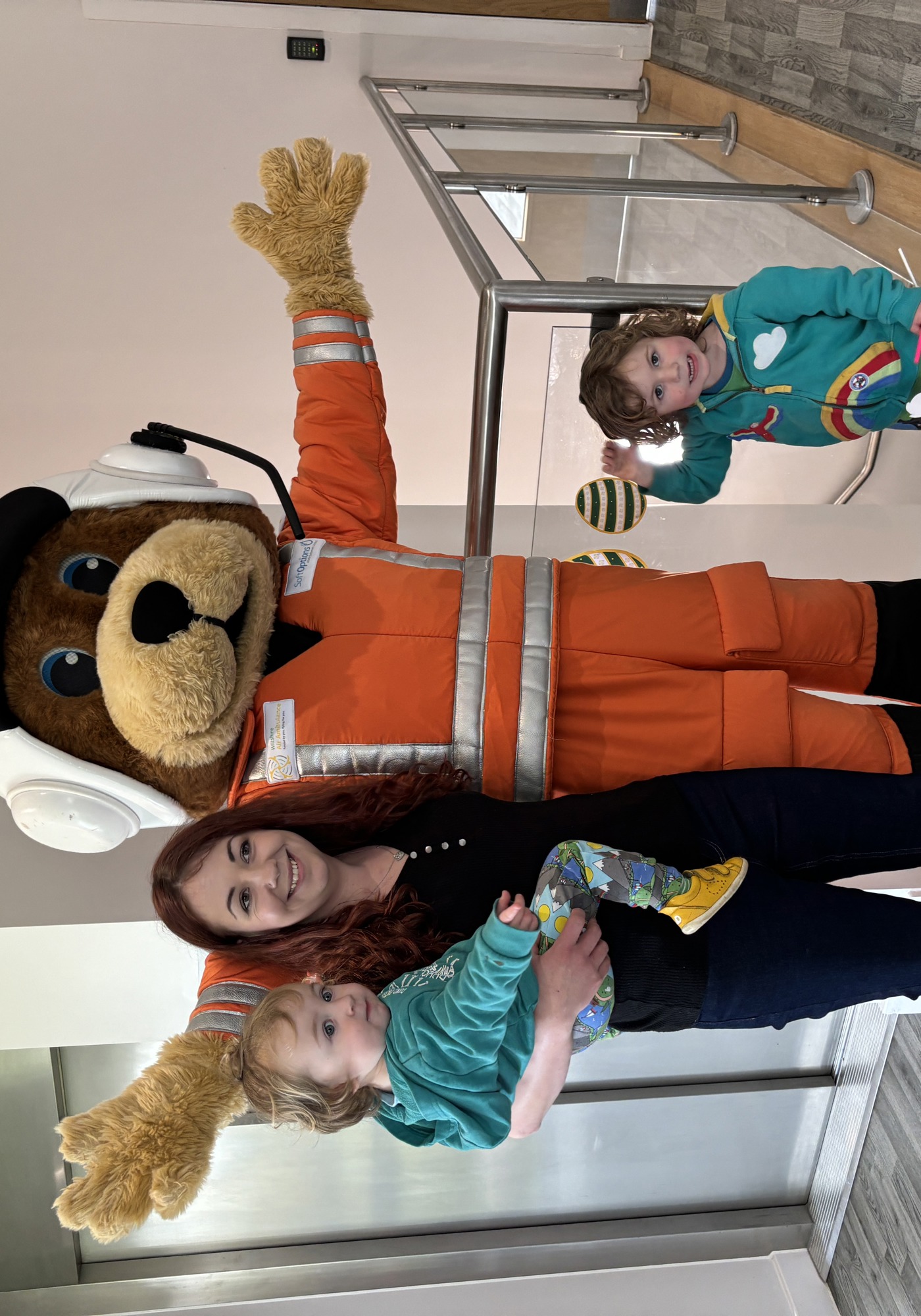 An orange paramedic bear mascot posting with an adult, holding a small child, and another child standing with them An orange paramedic bear mascot posting with an adult, holding a small child, and another child standing with them