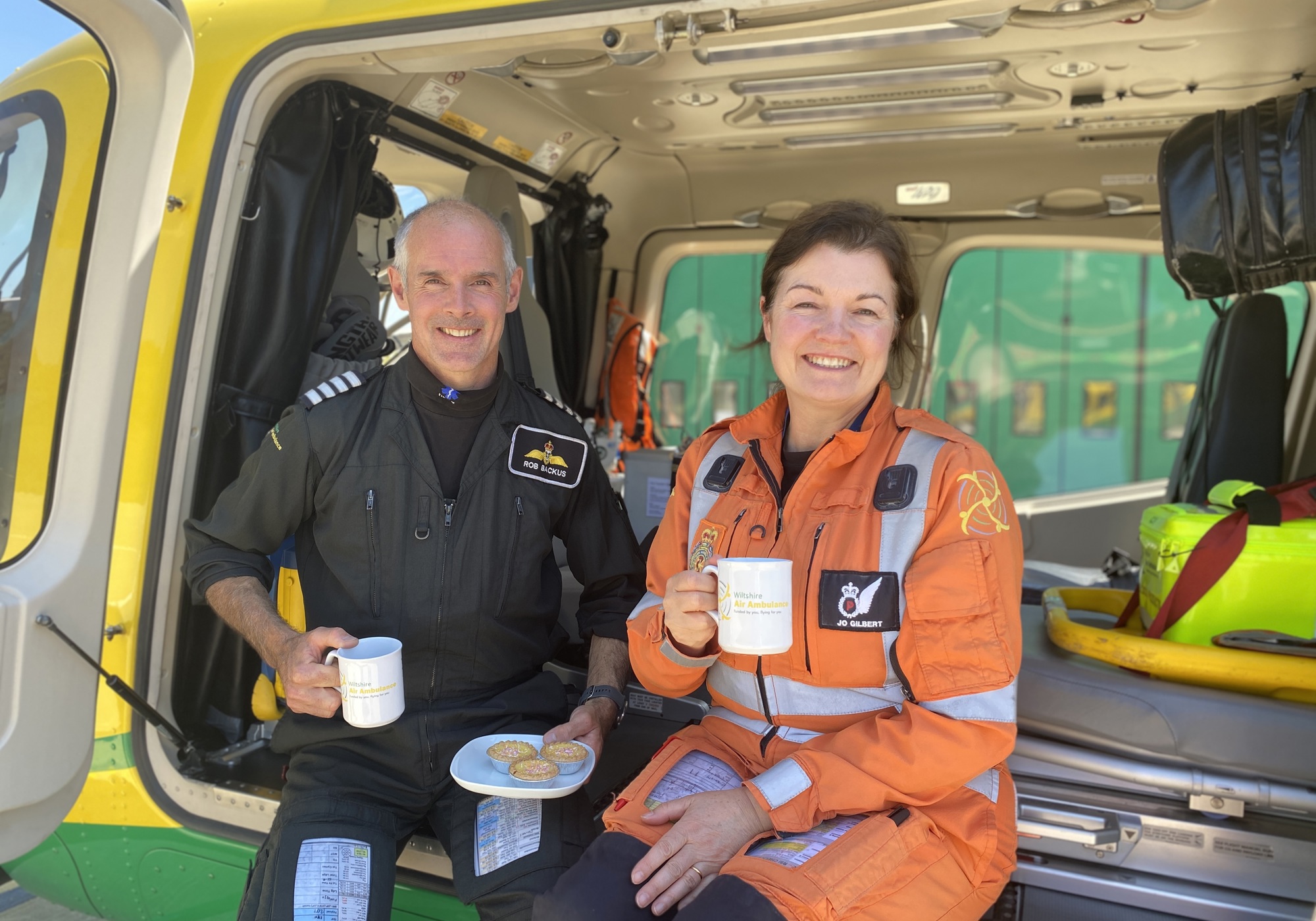 Pilot Rob and paramedic Jo posing with cakes and mugs whilst sat in Wiltshire Air Ambulance's helicopter